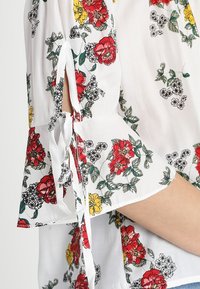Blommig blus i vit tyg, prydd med röda och gula blommor samt gröna blad. Har urklippta ärmar med knytband och en vida ärmdesign.