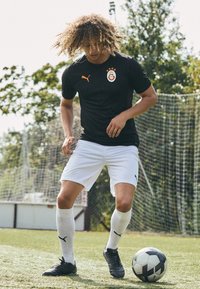 Maglietta da calcio nera con emblema arancione, pantaloni bianchi e scarpini neri. Giocando su erba artificiale con un pallone da calcio bianco e nero vicino.
