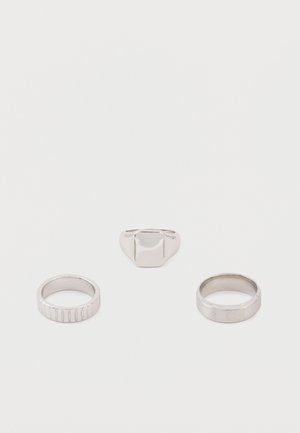 Τρία ασημένια δαχτυλίδια σε λευκή επιφάνεια: το ένα με χαραγμένες κάθετες γραμμές, ένα απλό δαχτυλίδι και ένα σινιέ δαχτυλίδι με επίπεδη ορθογώνια επιφάνεια.