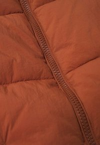 Gros plan d’un tissu matelassé couleur rouille avec une fermeture éclair en plastique visible et centrée, positionnée verticalement.