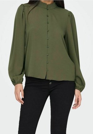 Mujer con blusa verde oliva abotonada con mangas largas abullonadas y pantalones negros ajustados sobre un fondo liso.