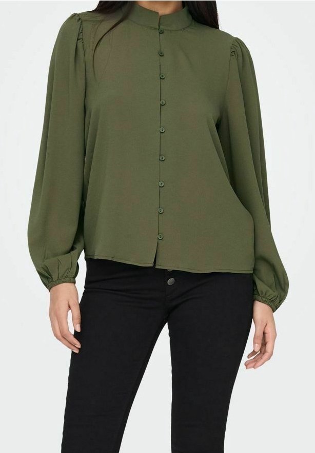 Femme portant un chemisier vert olive boutonné à manches longues bouffantes et un pantalon noir ajusté sur un fond uni.