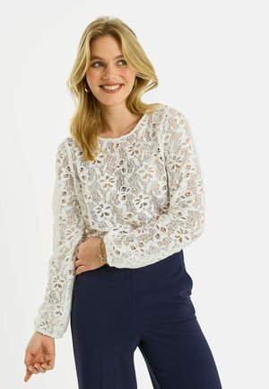 Mujer sonriente con cabello rubio, lleva una blusa blanca de encaje floral de manga larga y pantalones azules marino, posando con una mano en la cintura.