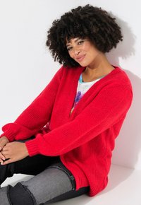 Cardigan rosso lavorato a maglia con scollo a V, spalle cadute e maniche lunghe. Abbinato a jeans grigi strappati. Sfondo bianco.
