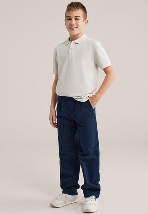 Tricou polo gri deschis, pantaloni bleumarin și adidași albi. Croială casual cu mâneci scurte, logo vizibil pe tricou și buzunare la pantaloni.