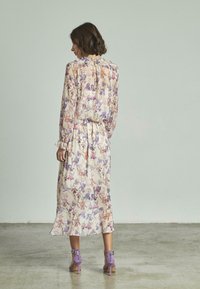 Robe midi florale à manches longues, fabriquée en tissu léger dans des couleurs pastel. Présente un col à volants et un ourlet évasé. Talons violets visibles.