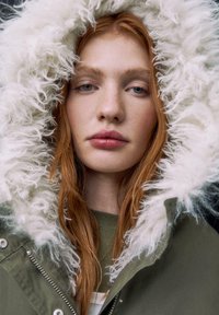 Grüne Parka mit einer flauschigen weißen Kunstfellkapuze. Glatte Stofftextur mit Reißverschluss und Druckknöpfen. Das Model hat rote Haare und ein natürliches Make-up.