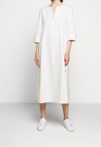 Bílá bavlněná šaty s výstřihem do V, tříčtvrtečními rukávy, bočními rozparky a rovný střih, doplněné bílými teniskami na neutrálním pozadí.