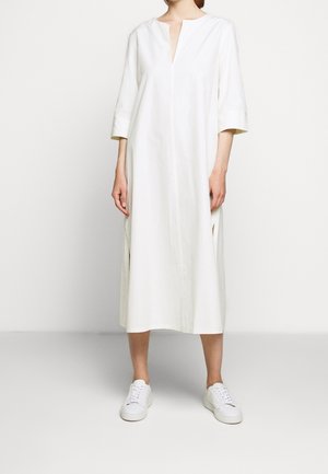 Robe en coton blanche avec un décolleté en V, des manches trois-quarts, des fentes sur les côtés et une coupe droite, associée à des baskets blanches sur un fond neutre.