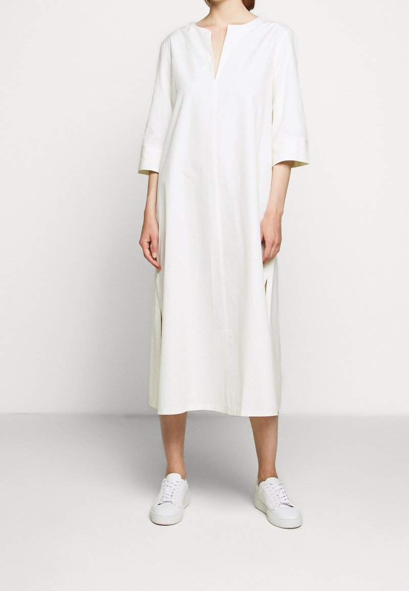 Bílá bavlněná šaty s výstřihem do V, tříčtvrtečními rukávy, bočními rozparky a rovný střih, doplněné bílými teniskami na neutrálním pozadí.