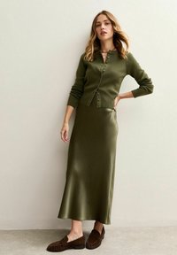 Femme portant un cardigan boutonné vert olive, une jupe midi en satin assortie et des mocassins en daim marron, se tenant devant un mur neutre.