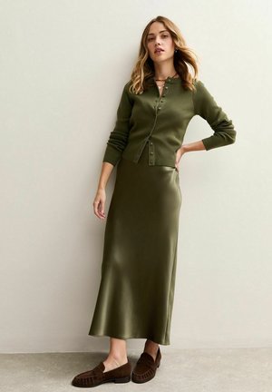 Woman wearing an olive green buttoned cardigan, matching satin midi skirt, and brown suede loafers, standing against a neutral wall.