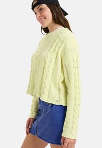 Junge Frau mit einem Lächeln, die einen hellgelben Zopfmusterpullover, einen blauen Jeansrock mit Reißverschluss vorne und eine schwarze Snapback-Cap, die rückwärts getragen wird, trägt.