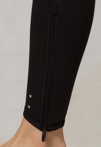 Czarne legginsy sportowe mają boczny zamek błyskawiczny i dwa odblaskowe kwadraty w pobliżu dołu. Tkanina ma gładką, elastyczną teksturę.