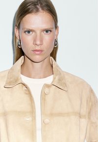 Beige suède jas met een kraag, voorzien van twee grote knopen en witte stiksels. Draagt over een lichtgekleurde blouse. Soepele textuur.