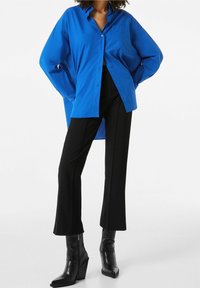 Camisa azul de botones y mangas largas, con un bolsillo; combinada con pantalones negros acampanados y botas de tacón negras con acabado texturizado.
