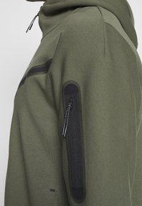 Olivgrön huvtröja med svart dragkedjeficka på ärmen, dragsko i huvan och diskreta axeldetaljer. Mjuk tygstruktur.