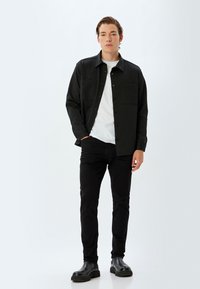 Camisa negra de botones con dos bolsillos en el pecho, llevada sobre una camiseta blanca. Jeans negros ajustados y botas de tobillo negras completan el conjunto.