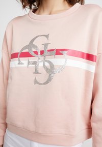 Vrouw draagt een lichtroze sweatshirt met rode en witte horizontale strepen en zilver glinsterende overlappende letters op de borst.