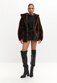 Frau steht und trägt eine braune Leopardenmuster Kunstpelzjacke, ein schwarzes langärmeliges Oberteil, schwarze Shorts und schwarze kniehohe Stiefel mit Absatz.