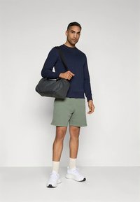 Tengerészkék pulóver, zöld rövidnadrág és fehér sportcipők. A modell sötétszürke sporttáskát visz. Simább textúrák, laza atlétikai stílus.