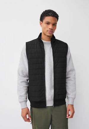 Giovane uomo che indossa un gilet trapuntato nero sopra una felpa grigio chiaro e pantaloni verde oliva, in piedi davanti a uno sfondo bianco semplice.