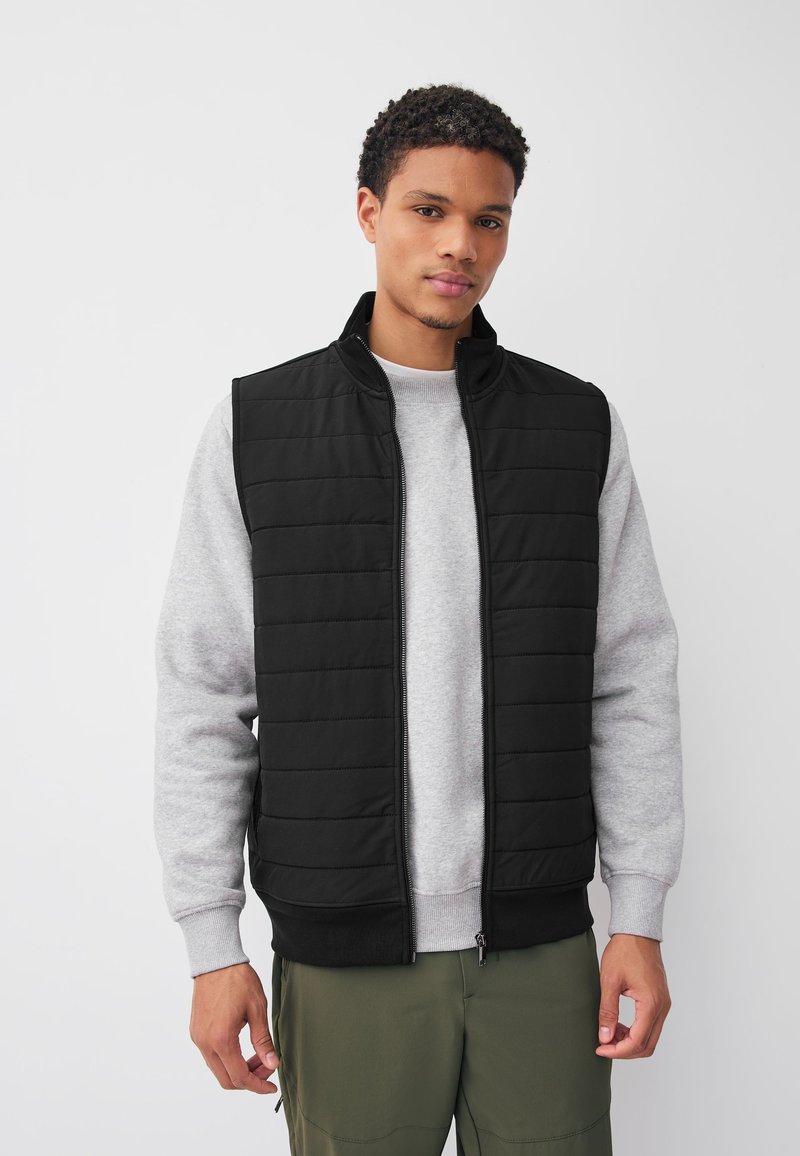 Jeune homme portant une veste matelassée noire sur un sweat-shirt gris clair et un pantalon vert olive, debout devant un fond blanc uni.