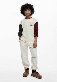 Cremefarbener Sweatshirt mit burgunderroten und schwarzen Ärmeln, mit einem gestickten Logo. Kombiniert mit cremefarbenen Jogginghosen mit roten Nähten und braunen Schuhen.