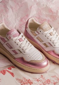 Paire de baskets en cuir blanc avec des touches métalliques roses et du daim beige, lacets blancs et bouts perforés sur papier floral froissé.