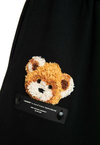 Tela negra con un parche de oso naranja texturizado y una etiqueta negra con texto blanco. Detalles suaves y de felpa en la cara y orejas del oso.