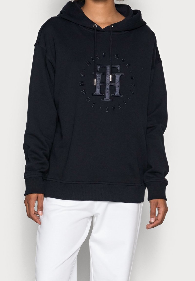 Black hoodie with a front logo and drawstring hood. Features ribbed cuffs and hem, made of a soft, structured fabric. Worn with white pants.