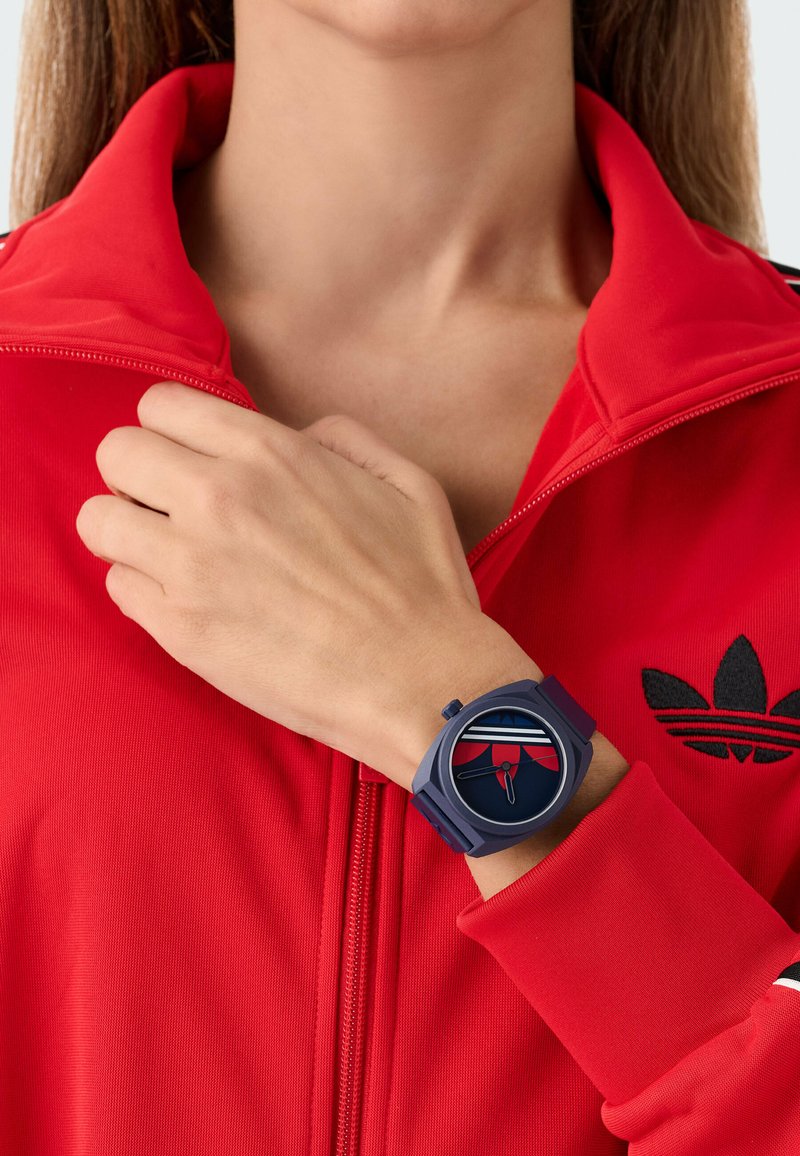 Wrist wearing a blue watch with a red, white, and blue patterned dial, against a red zip-up jacket with a black logo on the chest.