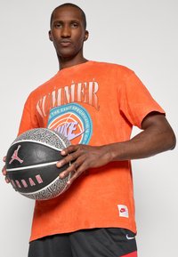 Orange bomulds t-shirt med grafisk print og rundt logo, parret med sorte shorts. Holder en struktureret basketball med røde og hvide detaljer.