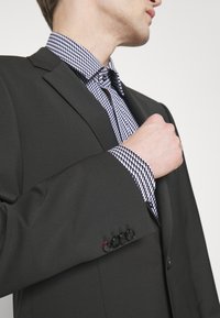 Abito nero con rever a punta, texture liscia, accentuato da un colletto di camicia a fantasia houndstooth e bottoni con dettagli rossi.