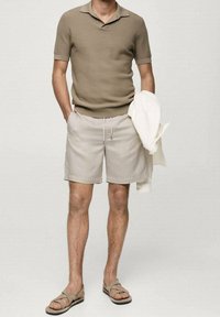 Polo en brun clair, tissu texturé ; short beige avec cordon de serrage ; sandales beige ; tenant une veste blanche.