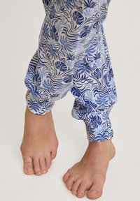 Blauwe en witte pyjamabroek met een bloemendessin, voorzien van elastische boorden en een relaxte pasvorm. Blote voeten zichtbaar aan de onderkant.