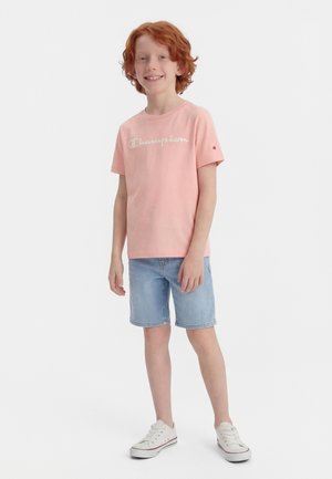 Raudonplaukis berniukas šypsosi, dėvi rožinį Champion marškinėlį, šviesiai mėlynus džinsinius šortus ir baltus sportinius batelius, stovėdamas ant vientisos baltos fono.