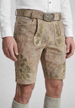 Man draagt traditionele beige leren lederhosen korte broek met groene borduursels, een riem met een dierlijke gesp, wit overhemd en gebreide sokken.