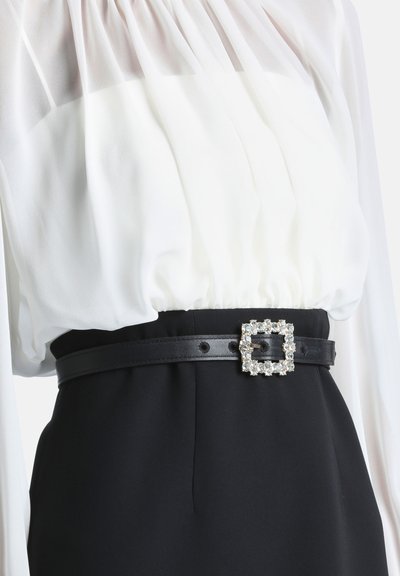 Ceinture en cuir noire avec boucle carrée ornée de strass portée sur un chemisier blanc et un pantalon noir à la taille d'une personne.