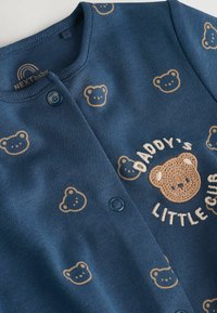 Mørkeblå bomuldsstrikjakke med bjørnemønster og broderet tekst "Daddy's Little Cub." Har to blå knapper og rund hals.