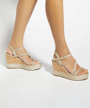Jambes portant des sandales compensées beige à brides avec semelle compensée en liège sur fond blanc uni.
