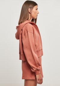 Kuttet hettegenser i støvete rosa, laget av mykt stoff. Har vide ermer og ribbede mansjetter, sammen med matchende shorts.