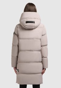 Párnázott, térd hosszúságú kabát világos bézs színben, nagy kapucnival, oldalsó zsebekkel és márkás felirattal a bal alsó oldalon. Simaság jellemzi.