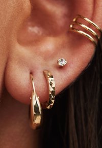 Goldene Creolen mit glänzender Oberfläche, begleitet von einem strukturierten goldenen Band und einem kleinen Diamantstecker, vor Hauthintergrund.