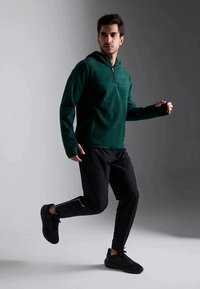 Pulôver de fleece verde floresta com fecho, de mangas compridas, combinado com calças de treino pretas e sapatilhas pretas, em fundo cinza.