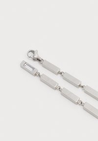 Braccialetto geometrico argentato con maglie rettangolari e una pietra trasparente rettangolare, chiuso con un gancio a moschettone. Texture liscia in tutto il bracciale.