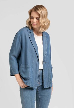 Donna con capelli biondi mossi che indossa un blazer di denim azzurro chiaro sopra una camicia bianca e jeans di denim abbinati, guarda verso il basso con le mani nelle tasche.