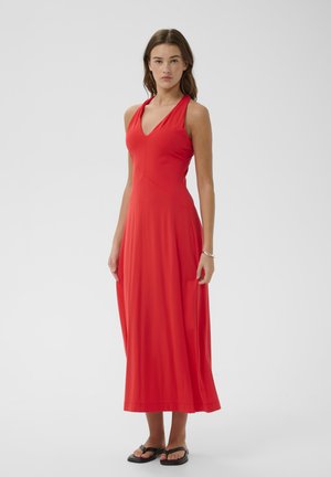 Mujer con vestido largo rojo sin mangas con escote en V y sandalias planas negras, de pie frente a un fondo blanco liso.