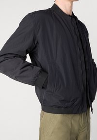 Veste bomber noire avec poignets et col côtelés. Présente une texture lisse et légère ainsi que des poches avant. Associée à un pantalon vert olive.