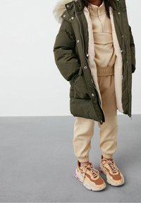 Manteau matelassé vert foncé avec capuche bordée de fourrure, haut beige superposé et joggeurs beiges. Chaussures de sport marron et rose complètent la tenue.